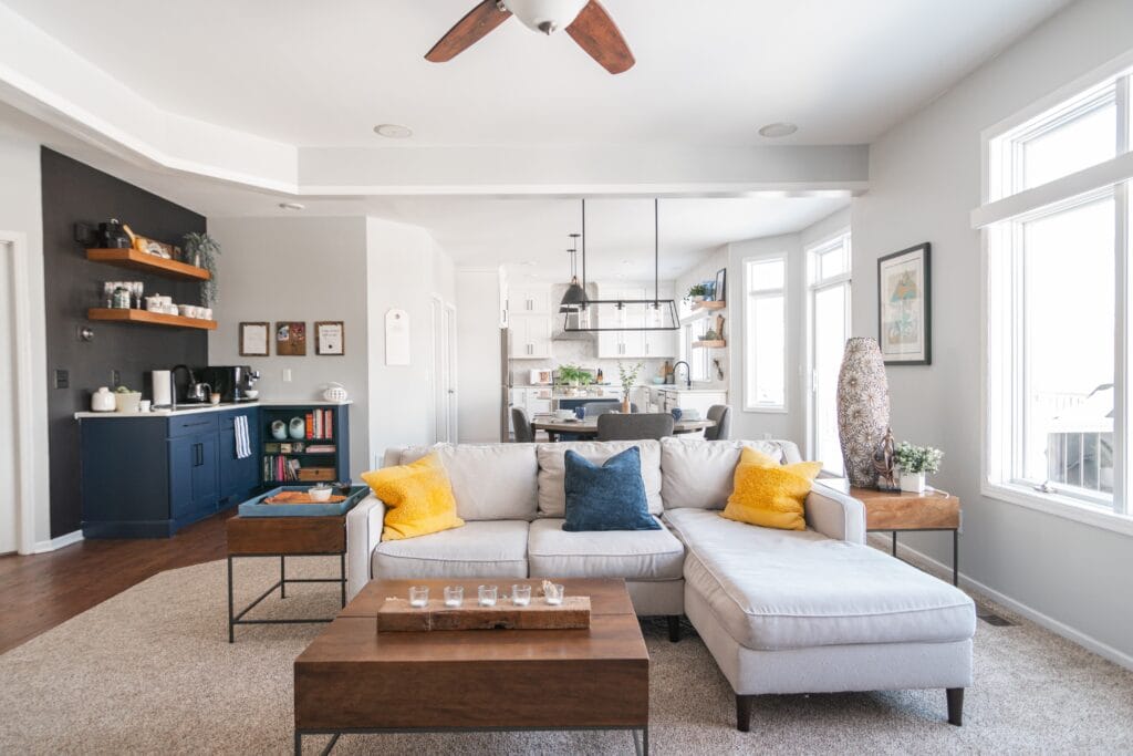 Living room with a kitchen in the background. Yellow pillows and white couch with ceiling fan.